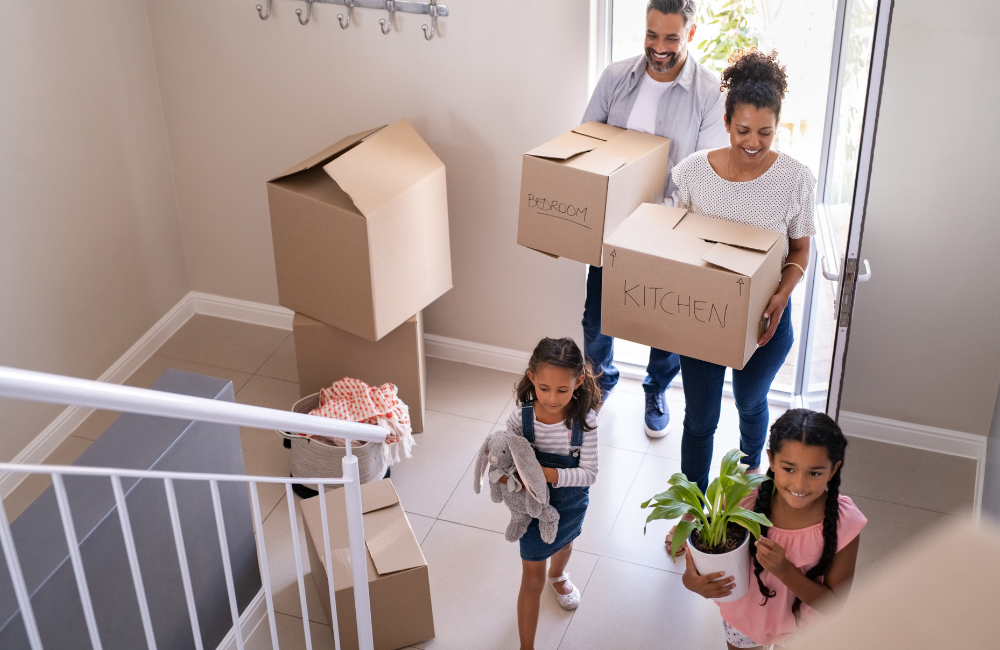 A family walking into their new home smiling. There are a couple of moving boxes inside. The mother and father are holding a moving box each while one daughter carries a small plant and the other carries plush bunny.