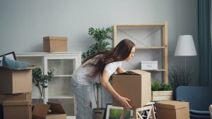 A woman placing a moving box down on a stool. She is surrounded by other belongings being packed to move.