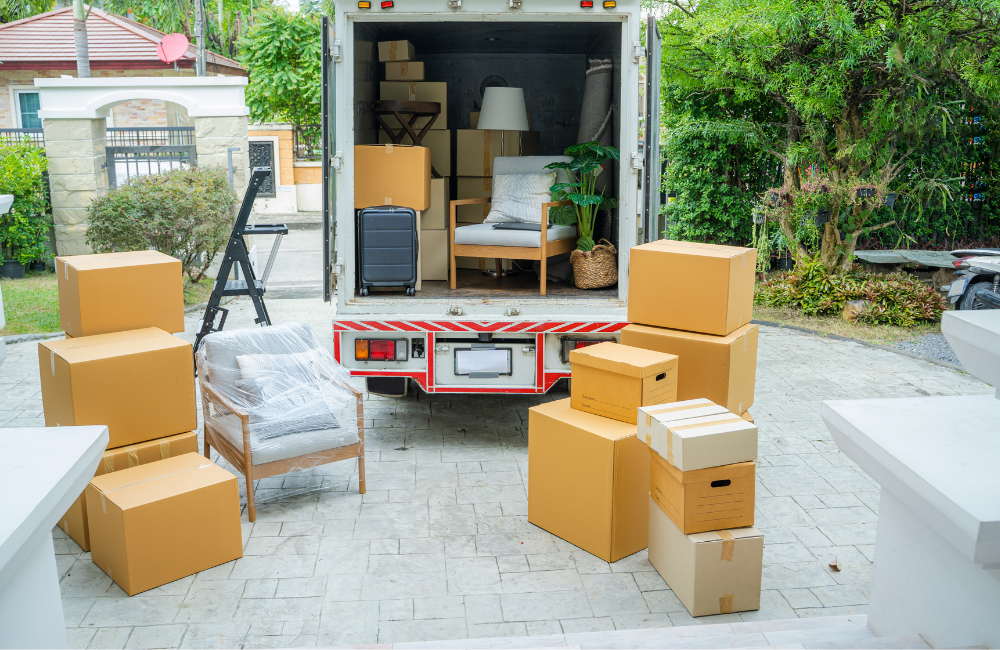 The back of a removalist truck in a driveway. The back doors are open showing household items inside and numerous other items around on the driveway. 