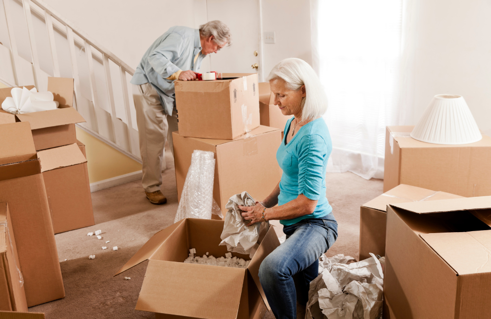 An older couple packing moving boxes in a lounge room. 