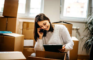 A woman on the phone surrounded by moving boxes and holding a clipboard. She is smiling and relaxed.