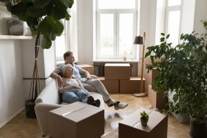 An older couple relaxing on a couch smiling with moving boxes around them in a lounge room.